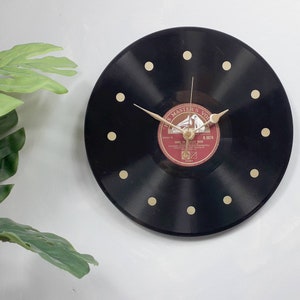 May include: A black vinyl record clock with gold-colored hands and hour markers. The center label is red and features the text "THE MASTER'S VOICE". The clock is mounted on a white wall, with green leaves in the background.