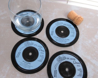 Può includere: Quattro sottobicchieri a forma di dischi di Michael Jackson con bordi neri e centri azzurri. Un sottobicchiere contiene un bicchiere d'acqua. Un tappo di sughero è vicino. I sottobicchieri presentano titoli di canzoni.