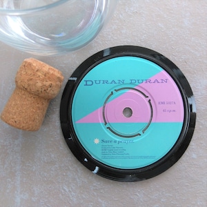 May include: A drinks coaster made from a vintage vinyl record, featuring a teal and pink design with the text "Duran Duran" and "Save a prayer." A cork bottle stopper and a glass bowl are also in the image.