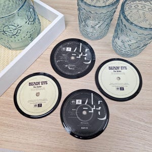 May include: Four round coasters with a vinyl record design. The coasters are black with white and beige labels, featuring text and graphics. Two blue patterned glasses and a white tray are also visible.