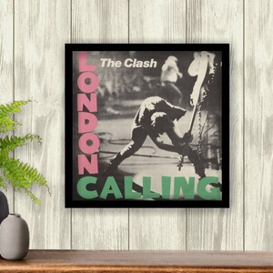 Op de afbeelding: Een ingelijste poster met de albumhoes van The Clash's "London Calling". De afbeelding is zwart-wit, met de bandnaam en albumtitel in roze en groen. De afbeelding toont een muzikant die een gitaar vernielt.