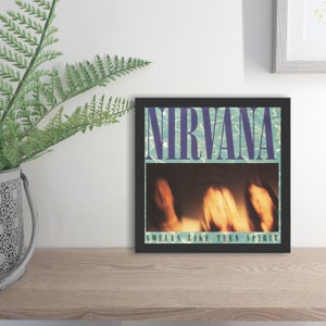 May include: A framed print of the Nirvana album cover for "Smells Like Teen Spirit." The artwork features the band's name in purple over a blue water background. Below, a blurred image of the band members is visible. The text "Smells Like Teen Spirit" is at the bottom.