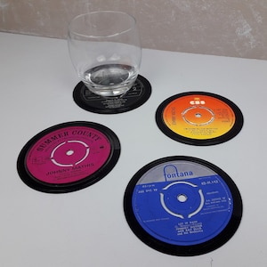 May include: Four vinyl record coasters with black rims. A glass sits on one. The record labels are in various colours, including pink, orange, and blue, with text such as "Johnny Mathis" and song titles.