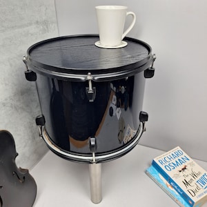 May include: A black drum converted into a side table. The table has a dark wood top and a single silver leg. A white mug sits on a coaster on the table. A book is visible on the floor.