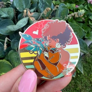 May include: A gold enamel pin featuring a woman with brown skin and curly brown hair holding a potted plant. The woman is wearing a gold earring. The background is a red, yellow, and green sunset.