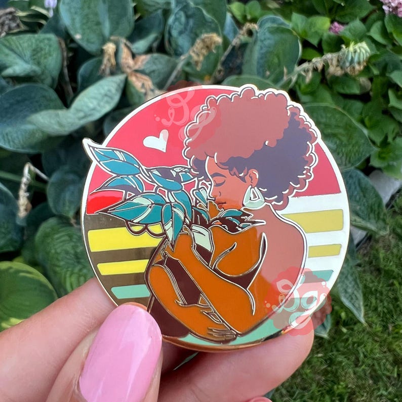 May include: A gold enamel pin featuring a woman with brown skin and curly brown hair holding a potted plant. The woman is wearing a gold earring. The background is a red, yellow, and green sunset.