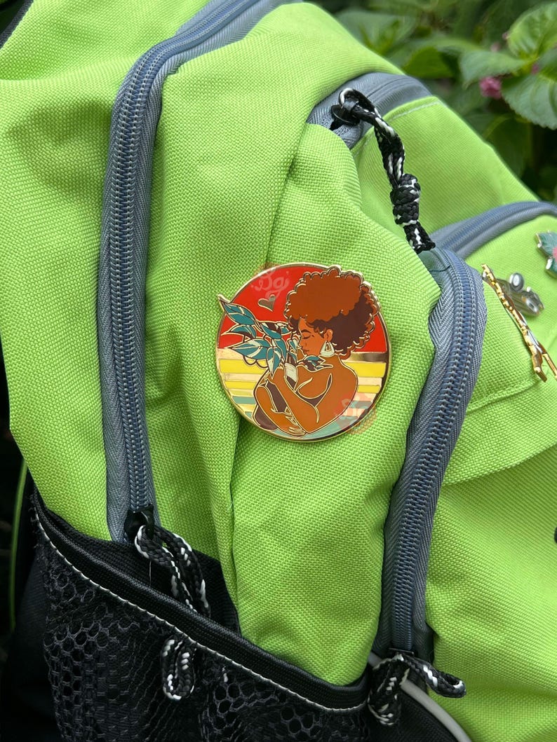 May include: A gold-toned enamel pin featuring a woman with brown hair and a plant. The woman is wearing a gold hoop earring and a gold necklace. The background is a sunset with red, orange, and yellow colors. The pin is attached to a green backpack.