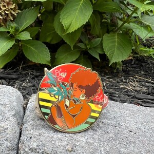 May include: A round enamel pin featuring a woman with red hair holding a potted plant. The woman is wearing a brown dress and has a large hoop earring. The background is a yellow, orange, and red striped pattern with the word "Soji" in white.