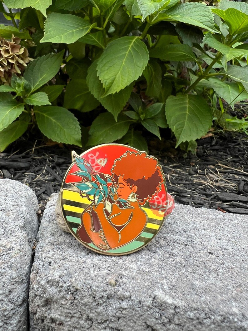May include: A round enamel pin featuring a woman with red hair holding a potted plant. The woman is wearing a brown dress and has a large hoop earring. The background is a yellow, orange, and red striped pattern with the word "Soji" in white.