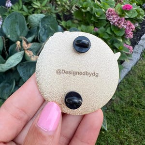 May include: Gold-colored round pin with a black and white design. The back of the pin has two black metal clasps and the text "@Designedbydg".