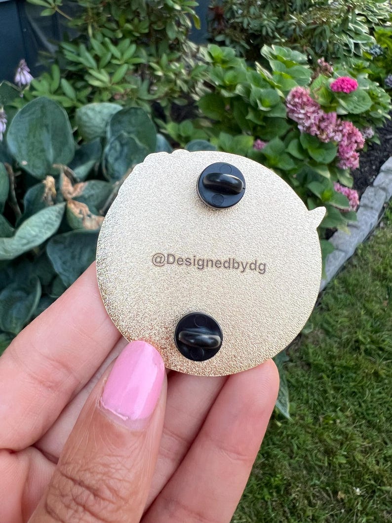 May include: Gold-colored round pin with a black and white design. The back of the pin has two black metal clasps and the text "@Designedbydg".