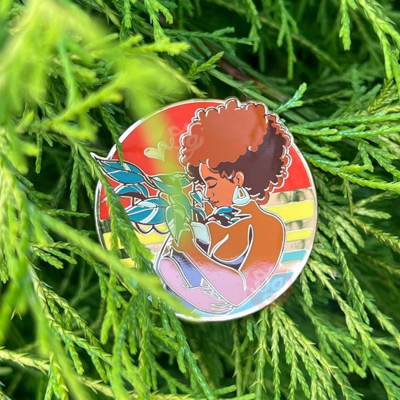 May include: A colorful enamel pin featuring a woman with brown hair holding a plant. The background is a sunset with a rainbow stripe.