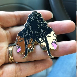 May include: Gold-tone enamel pin featuring two black and white cartoon faces with pink tongues sticking out. The faces are joined together by a black and white cartoon hairstyle.