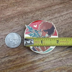 May include: A gold-toned enamel pin featuring a woman with brown skin and curly brown hair holding a green plant. The woman is wearing a red and white patterned dress and a heart-shaped earring. The pin is approximately 1 inch in diameter.