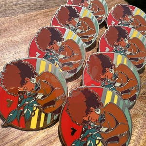 May include: A set of nine enamel pins featuring a woman with an afro holding a plant. The pins are round and have a red, yellow, and green striped background. The woman is depicted in a brown skin tone and is wearing a brown shirt. The plant is green and has white leaves.