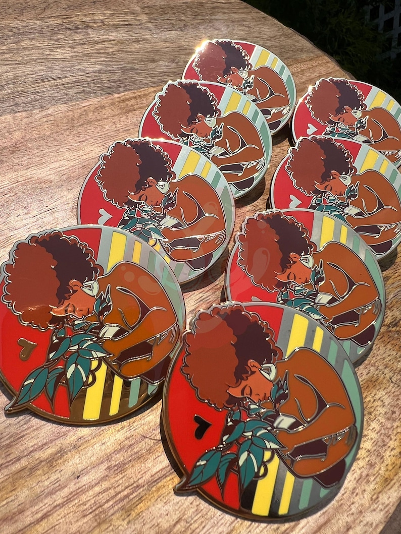 May include: A set of nine enamel pins featuring a woman with an afro holding a plant. The pins are round and have a red, yellow, and green striped background. The woman is depicted in a brown skin tone and is wearing a brown shirt. The plant is green and has white leaves.