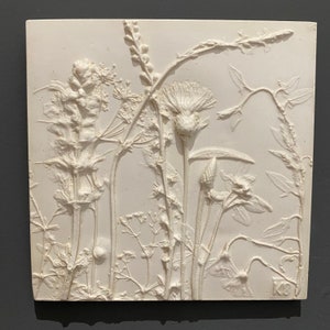 May include: A white ceramic relief sculpture of a group of wildflowers. The sculpture is in a square frame and has a textured surface.