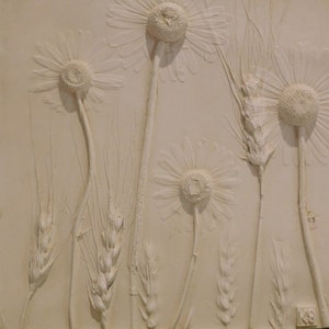 May include: A white plaster relief sculpture of daisies and wheat stalks. The sculpture is on a white background and has a signature in the lower right corner.