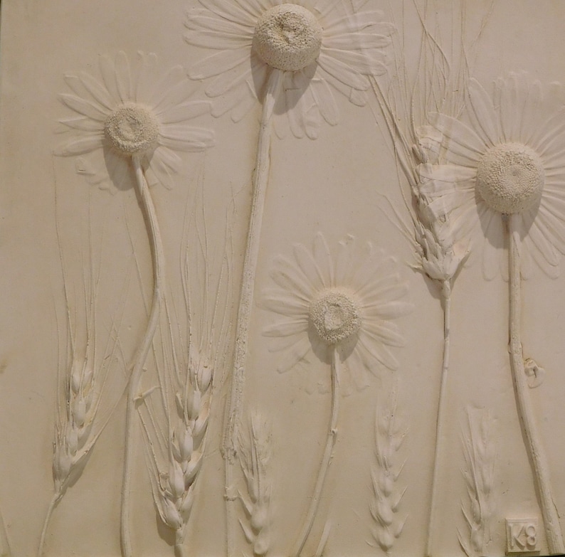 May include: A white plaster relief sculpture of daisies and wheat stalks. The sculpture is on a white background and has a signature in the lower right corner.