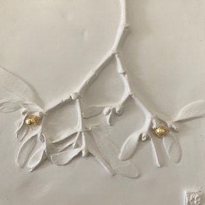 May include: A white relief sculpture of a mistletoe branch with leaves and berries. The berries are accented with gold leaf. The artwork is mounted on a white background, creating a monochromatic and textured aesthetic.