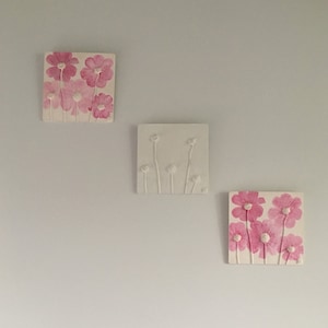 May include: Three square wall decor pieces with pink and white floral designs. The top piece features pink flowers with white centers, the middle piece features white flowers with white stems, and the bottom piece features pink flowers with white centers.