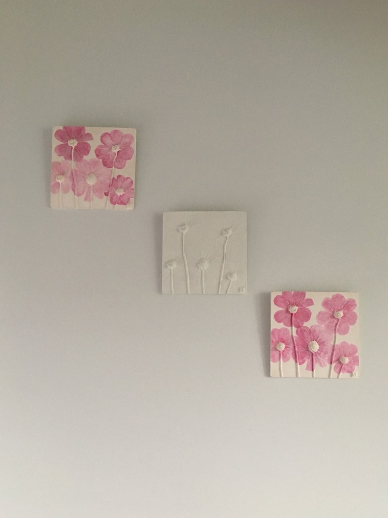 May include: Three square wall decor pieces with pink and white floral designs. The top piece features pink flowers with white centers, the middle piece features white flowers with white stems, and the bottom piece features pink flowers with white centers.