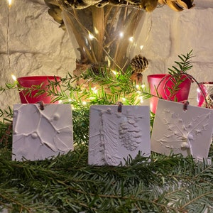 Könnte beinhalten: Drei weiße Keramik-Anhänger mit naturinspirierten Designs. Jeder Anhänger zeigt eine andere Pflanze oder einen Ast, darunter eine Kiefernzapfen, ein Blatt und ein Zweig. Die Anhänger hängen an roter und weißer Schnur.