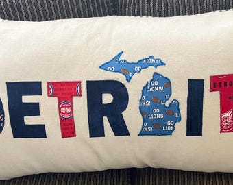May include: Cream-colored rectangular pillow with the word "DETROIT" in navy blue, red, and patterned fabric letters. The letter "D" has a Detroit Tigers logo, and the letter "I" has a Michigan outline with "GO LIONS!" text.