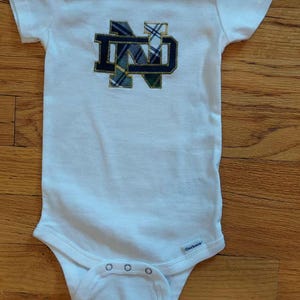 May include: White baby onesie with a navy blue, gold, and green embroidered "ND" logo. The onesie has short sleeves and snap closures at the bottom. The brand name "Gerber" is printed on the garment.