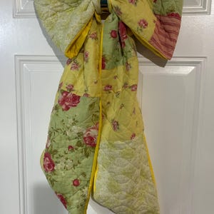 May include: A large decorative bow made from quilted fabric featuring a patchwork design. The fabric displays a mix of yellow, light green, and pink floral patterns, including roses and stripes. The bow has a bright yellow trim along the edges.