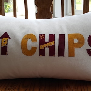 May include: White pillow with the word "CHIPS" spelled out in gold and maroon fabric. The letters are made from different pieces of fabric, including a small box with the word "CHIPPEWA" on it. There is a flame and an arrow pointing up on the left side of the pillow.