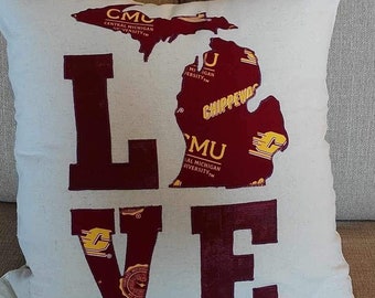 May include: Cream-colored pillow with the word "LOVE" in maroon block letters. The letter "O" is replaced by a maroon outline of the state of Michigan, with university names and logos in the center.