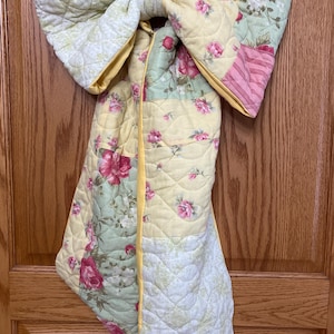 Peut inclure: Un grand nœud décoratif fabriqué en tissu matelassé avec un motif patchwork. Le textile présente un imprimé floral avec des roses roses, du feuillage vert et des accents jaunes, fini par un liseré en tissu jaune sur les bords.