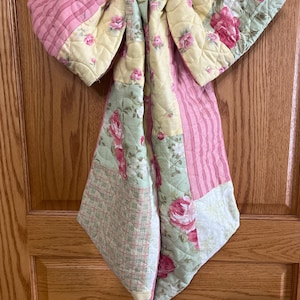 May include: A quilted fabric bow featuring a patchwork design with floral patterns in pink, yellow, and sage green, alongside sections of pink striped fabric and a small green and white checkered print, all stitched with a diamond quilting pattern.