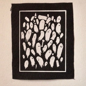 Pode incluir: Um patch de tecido preto com uma borda branca apresentando um padrão de figuras de fantasmas sorridentes brancos de vários tamanhos. Alguns fantasmas estão segurando objetos. O patch tem uma borda ligeiramente desfiada.