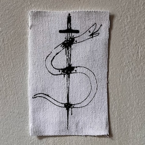 Könnte beinhalten: Ein gestickter Patch in Schwarzweiß, der eine Schlange zeigt, die sich um ein Schwert windet. Die Schlange ist stilisiert, fast cartoonartig, im Design. Das Schwert hat einen kreuzförmigen Griff und tropft mit schwarzer Tinte.