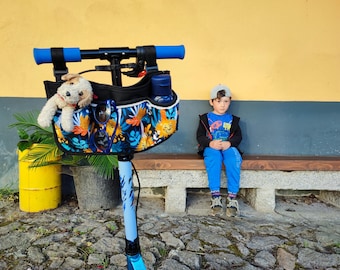 May include: A blue and black scooter with a floral print bag, a stuffed dog, and a water bottle. The scooter has blue handlebars and a blue stem. A young child in blue pants and a black jacket sits on a bench in the background.