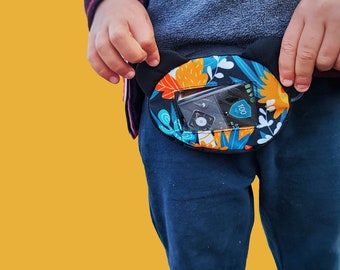 May include: A child wearing blue jeans and a black and blue jacket with a colorful floral print insulin pump pouch attached to their waist. The pouch has a clear window showing the insulin pump with a digital display reading "100".
