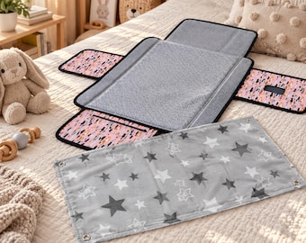 May include: A portable baby changing mat featuring a gray textured center panel with black trim and side flaps decorated with a pink, black, and white abstract pattern. A separate rectangular gray cloth with white and dark gray star prints is included.