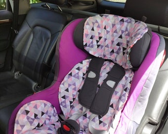 May include: A child safety seat featuring a vibrant purple frame and a removable fabric cover with a geometric pattern in shades of pink, white, black, and lavender. The seat includes dark gray shoulder harness pads and a central buckle system.