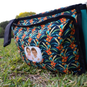 May include: A rectangular diaper bag with a vibrant floral pattern in navy, orange, and green. The bag features a black handle and trim, a teal side panel, and a small photo window with two baby pictures.