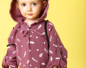 May include: A young child wearing a pink hooded onesie with a white feather pattern. The onesie has bear ears on the hood. The child is standing in front of a yellow background.