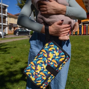 May include: A navy blue and black baby changing pad with a colorful pattern of yellow bananas and orange leaves. The pad has a black handle and a snap closure. The background shows a person holding a baby.