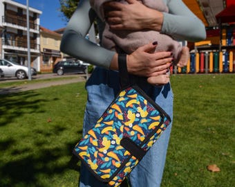 May include: A rectangular portable changing mat featuring a dark blue background with a vibrant pattern of yellow bananas, orange carrots, and green leaves. The mat includes a black fabric wrist strap and a black trim with a secure snap closure.