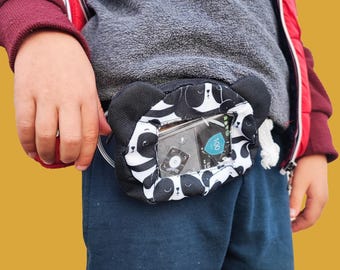May include: A black and white panda print fabric pouch with a clear plastic window. The pouch is worn on a person's waist and has a small device inside the window. The device has a screen that reads "100".