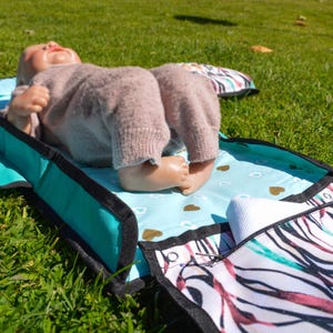 May include: A baby doll wearing a light brown outfit rests on a teal and patterned changing pad. The pad has a blue interior with gold heart shapes and a patterned border with black trim. The scene is set on green grass.