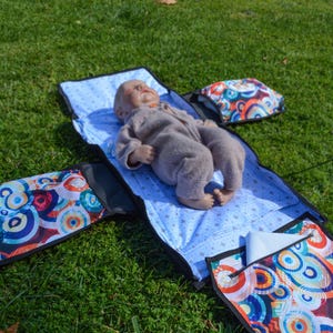 May include: A baby changing station with a blue patterned changing pad and colorful storage compartments. The changing pad is open, with a baby doll lying on it. The storage compartments have a colorful circular design.