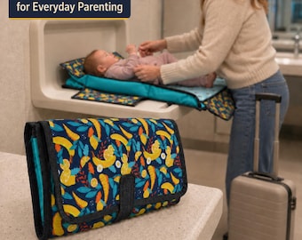 May include: A folded rectangular diaper clutch featuring a dark blue fabric with a vibrant pattern of yellow bananas, orange citrus slices, and teal leaves. The clutch has a black fabric trim and a central closure strap. Text reads: Compact Diaper Clutch for Everyday Parenting.