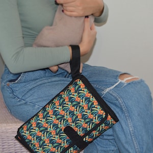 May include: A black changing pad with a colorful floral pattern featuring orange, yellow, and green leaves. The pad has a black handle and a black closure strap. The pad is folded and appears to be new.