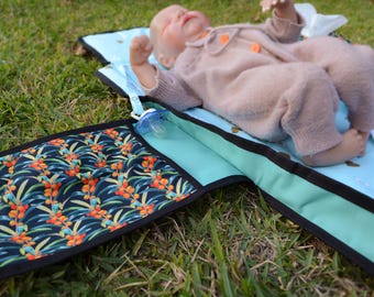May include: A portable baby changing mat featuring a light blue surface with a white pattern and a black-trimmed pocket decorated with a dark blue, green, and orange sea buckthorn berry print. A doll in a beige knit onesie lies on the mat.